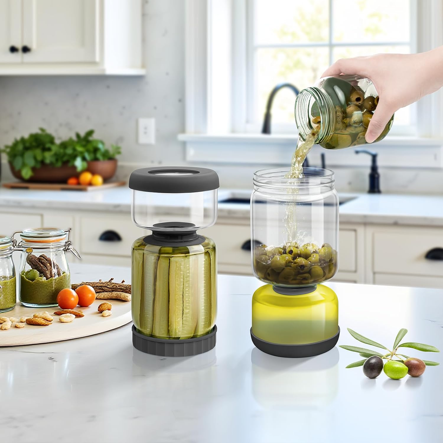 Two-part glass storage jar on a kitchen counter holding pickle spears above a lower chamber of drained brine, beside a matching jar of olives