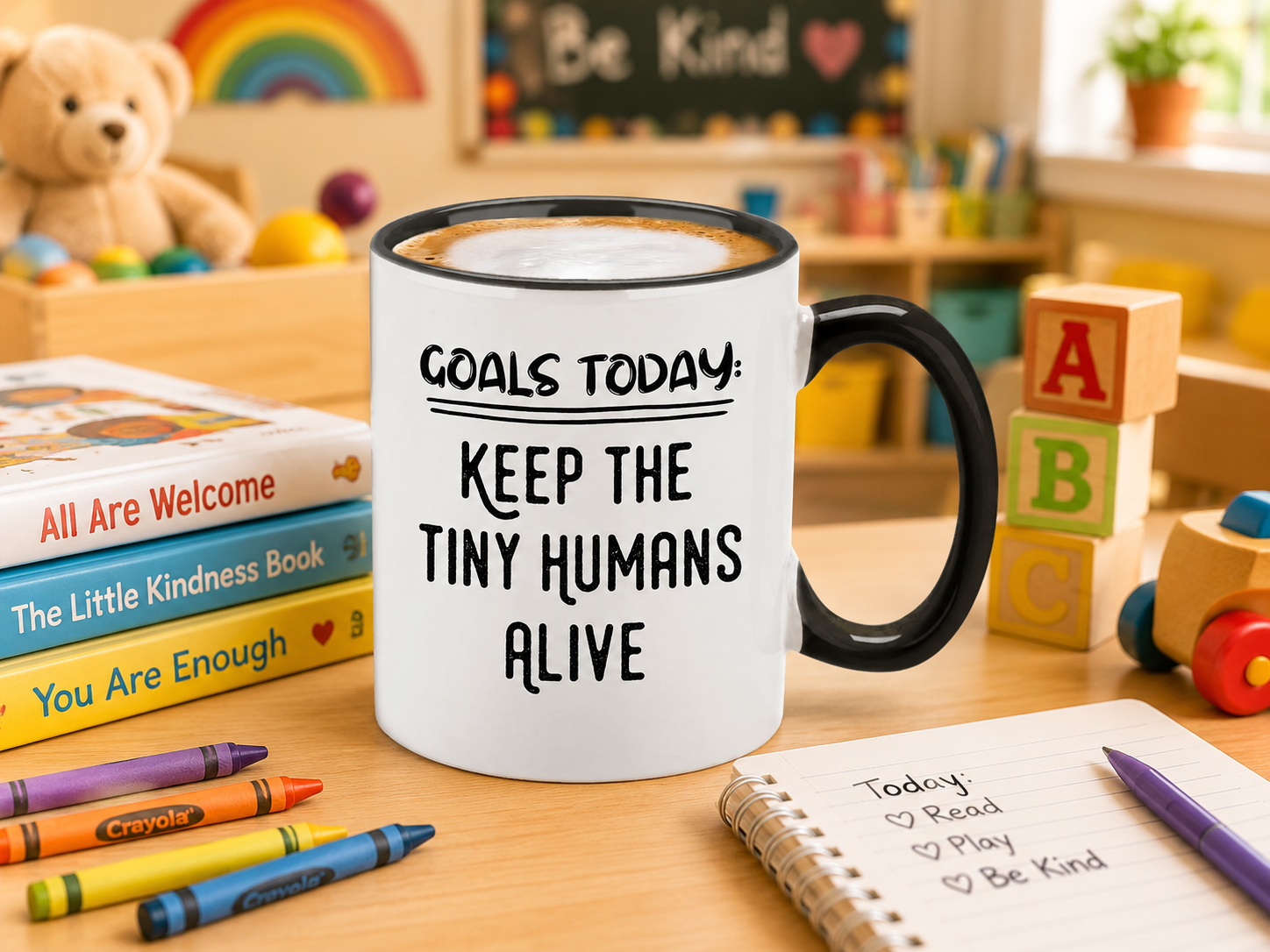 White ceramic coffee mug with black rim and handle and bold black 'Goals Today: Keep The Tiny Humans Alive' lettering styled in a bright playroom with picture books, wooden blocks, and crayons