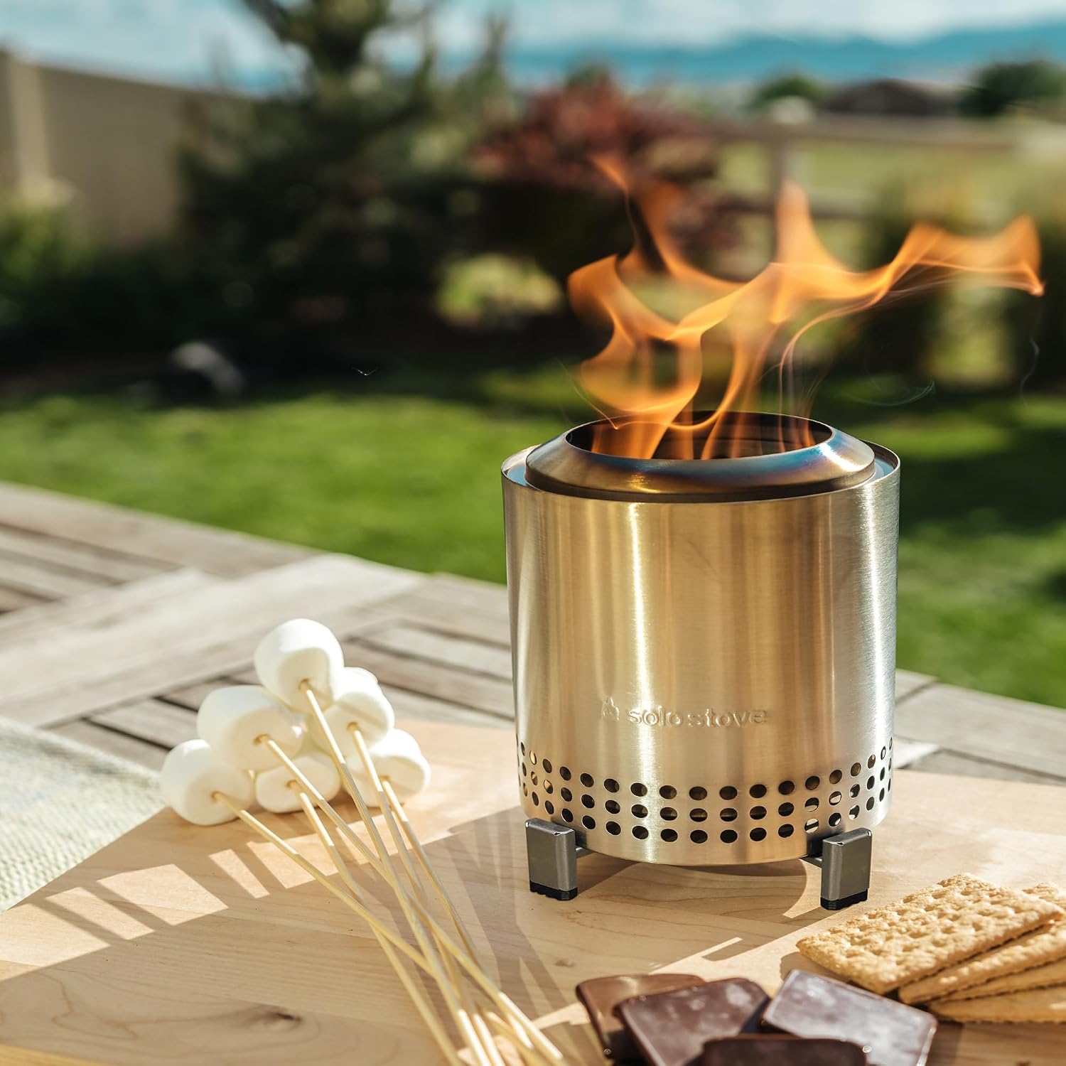 Stainless steel smokeless fire pit burning on a wooden outdoor table beside marshmallow skewers, graham crackers and chocolate