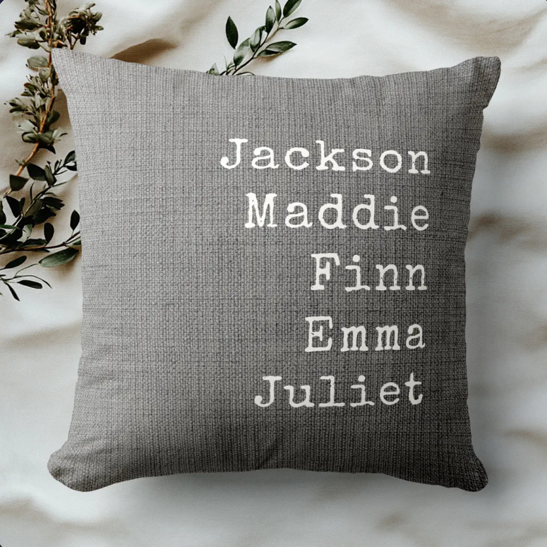 Grey woven farmhouse throw pillow printed with the names Jackson, Maddie, Finn, Emma and Juliet in cream typewriter lettering, styled with a sprig of greenery