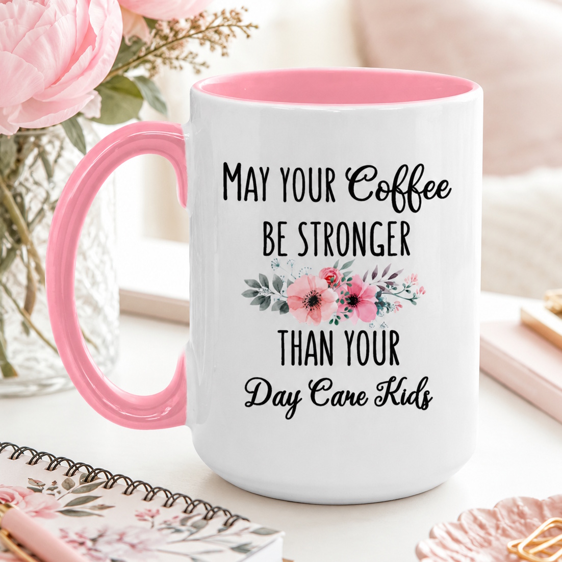 Funny white ceramic 'May Your Coffee Be Stronger Than Your Day Care Kids' pink-handled coffee mug gift for daycare teachers with pink floral accents styled beside peonies