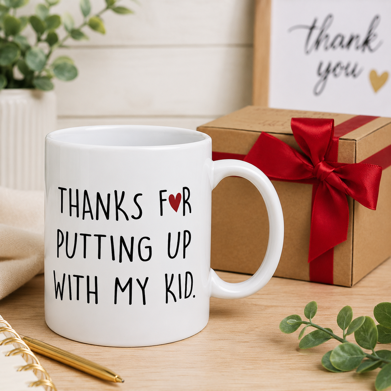 Funny white ceramic 'Thanks For Putting Up With My Kid' daycare teacher thank-you coffee mug with small red heart, styled beside a kraft gift box with red satin ribbon