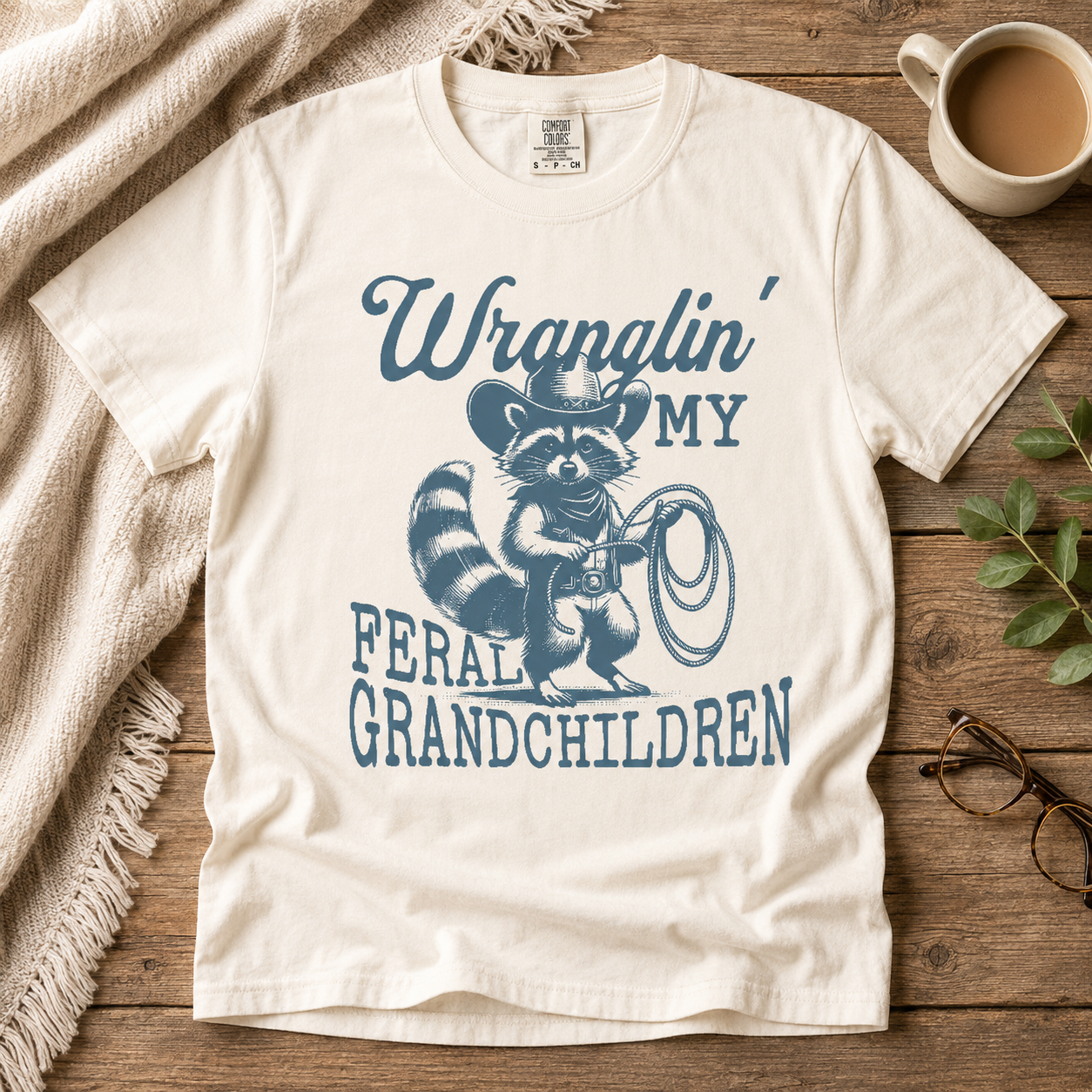 Cream cotton t-shirt printed in slate blue with a lasso-holding raccoon in a cowboy hat and the words Wranglin' My Feral Grandchildren