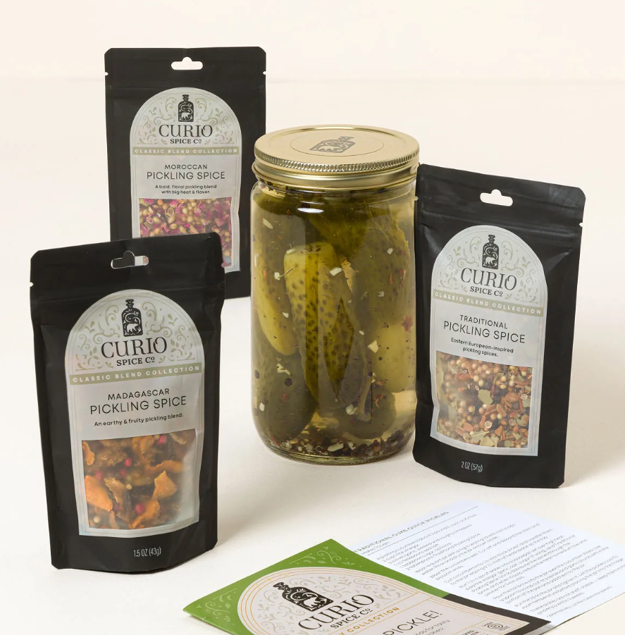Three pouches of Curio Spice Co. pickling blends arranged around a mason jar of pickled cucumbers with recipe cards in front