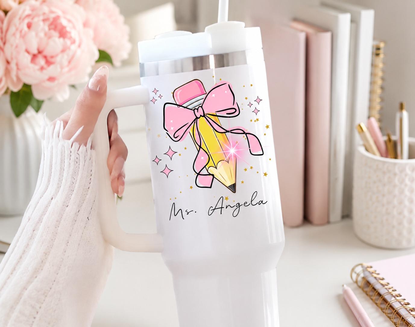 White personalized 40 oz teacher tumbler travel mug with pink bow and pencil design and hand-lettered 'Ms. Angela' name, styled beside pink peonies and pastel books
