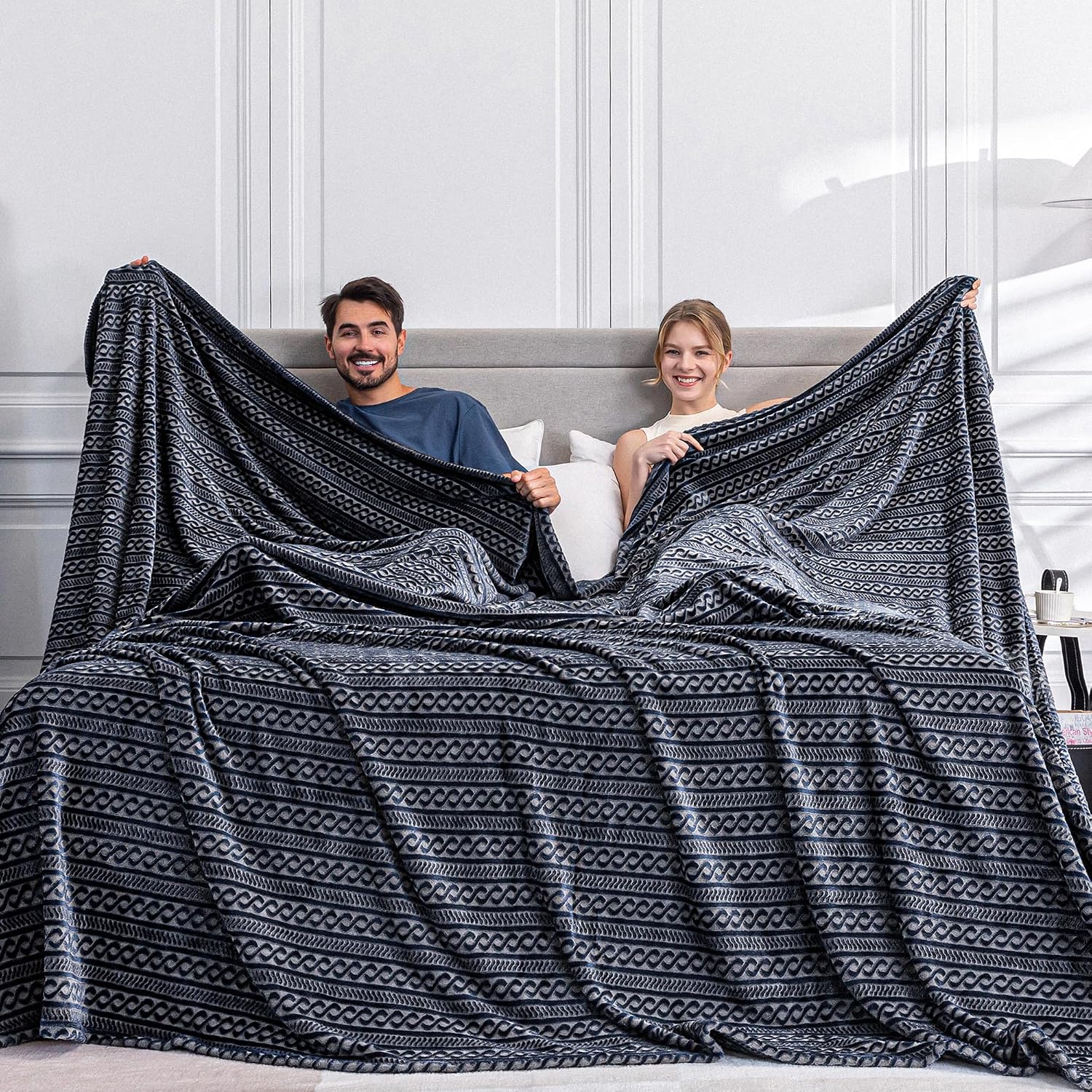 Extra-large navy and grey cable-pattern plush blanket spread across a bed, held open by two people
