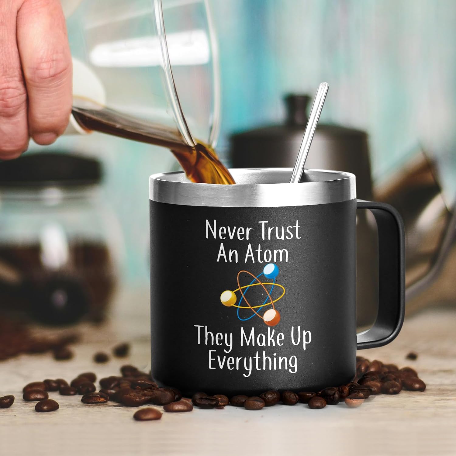 Matte black insulated science teacher travel mug with 'Never Trust An Atom They Make Up Everything' quote and colorful atom illustration, styled with fresh coffee being poured beside scattered coffee beans