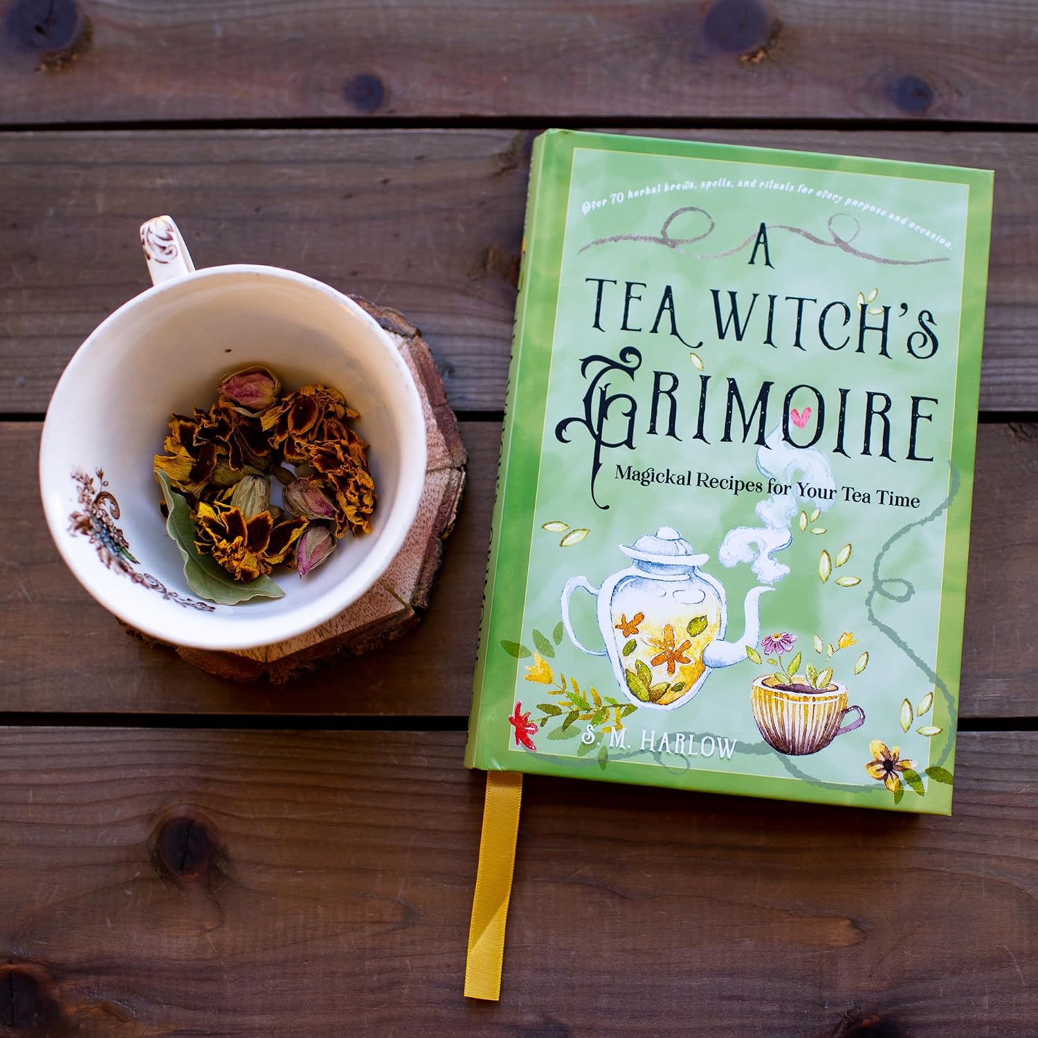 Hardcover copy of A Tea Witch's Grimoire with a green illustrated cover beside a teacup of dried rose buds and herbs on a wooden table