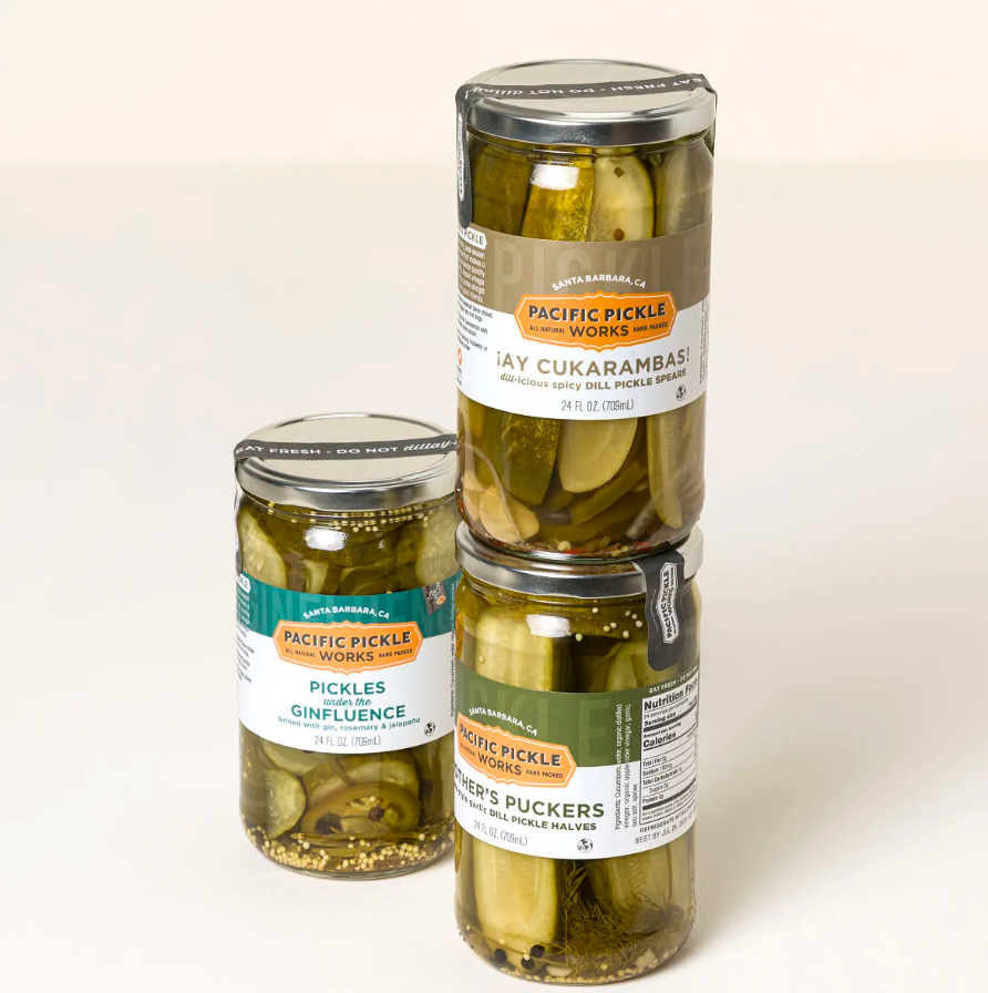 Three tall glass jars of handcrafted dill pickle spears and halves with printed labels, stacked against a pale background