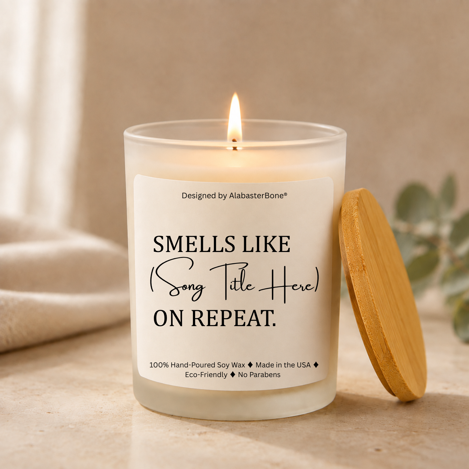 Frosted glass soy candle with a bamboo lid and a cream label reading Smells Like (Song Title Here) On Repeat, styled beside eucalyptus and a folded towel