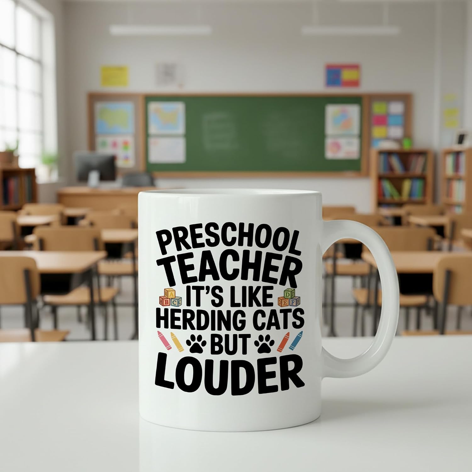 White ceramic preschool teacher coffee mug with bold black lettering 'Preschool Teacher It's Like Herding Cats But Louder' surrounded by playful crayon and ABC block illustrations
