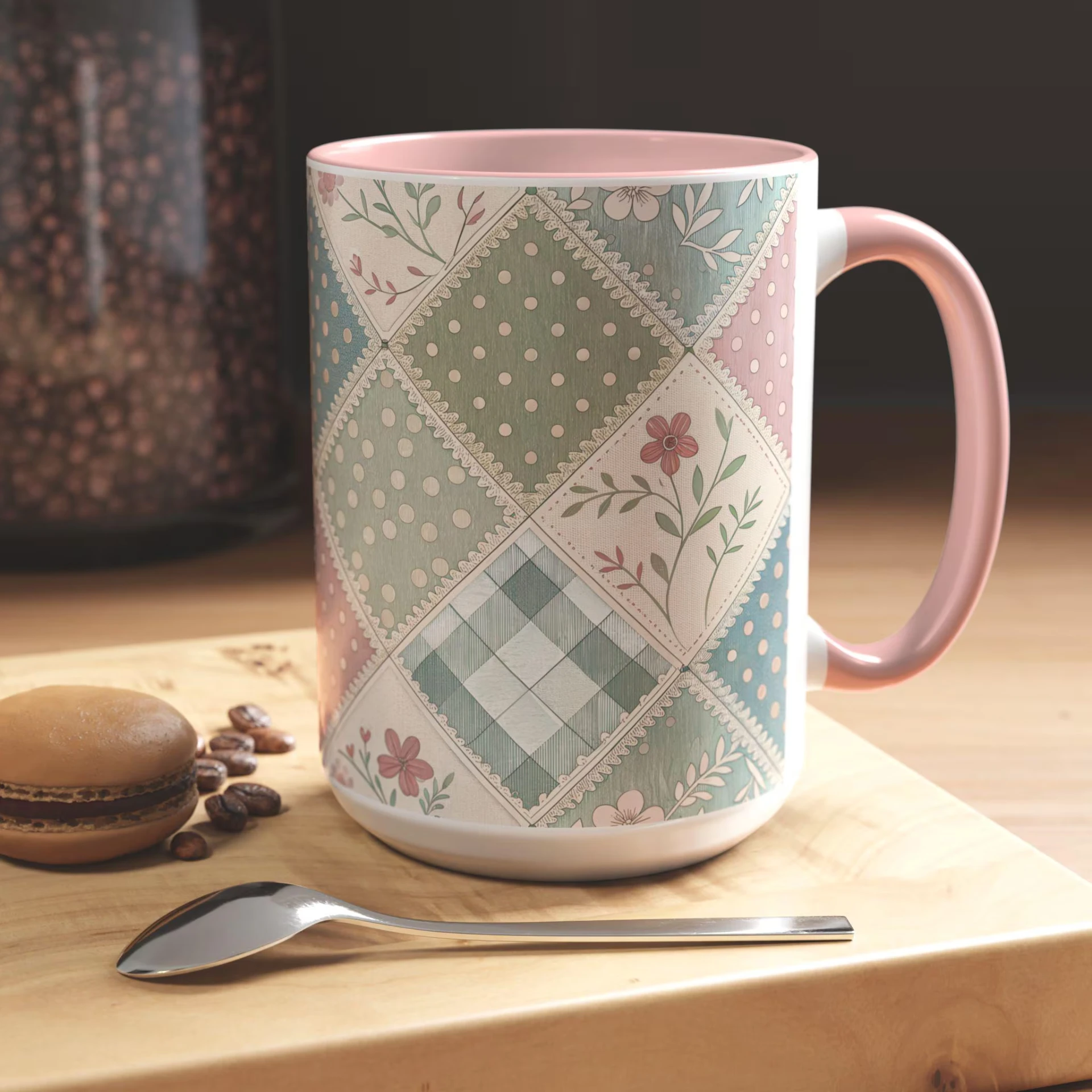 Pastel patchwork quilt-pattern mug with pink handle and interior on a wooden tray beside a macaron and coffee beans