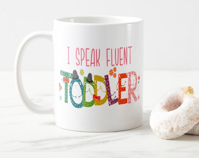 Playful white ceramic 'I Speak Fluent Toddler' daycare teacher coffee mug with colorful patterned monster letters styled on a marble surface with a powdered donut