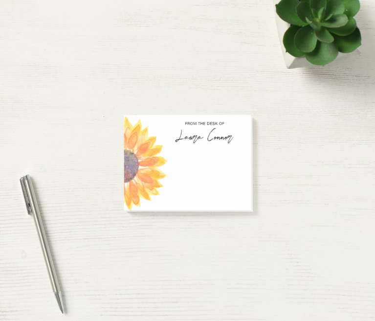 White personalized sticky note pad printed with a watercolor sunflower and the words From the Desk of Laura Connor, beside a silver pen and a small succulent